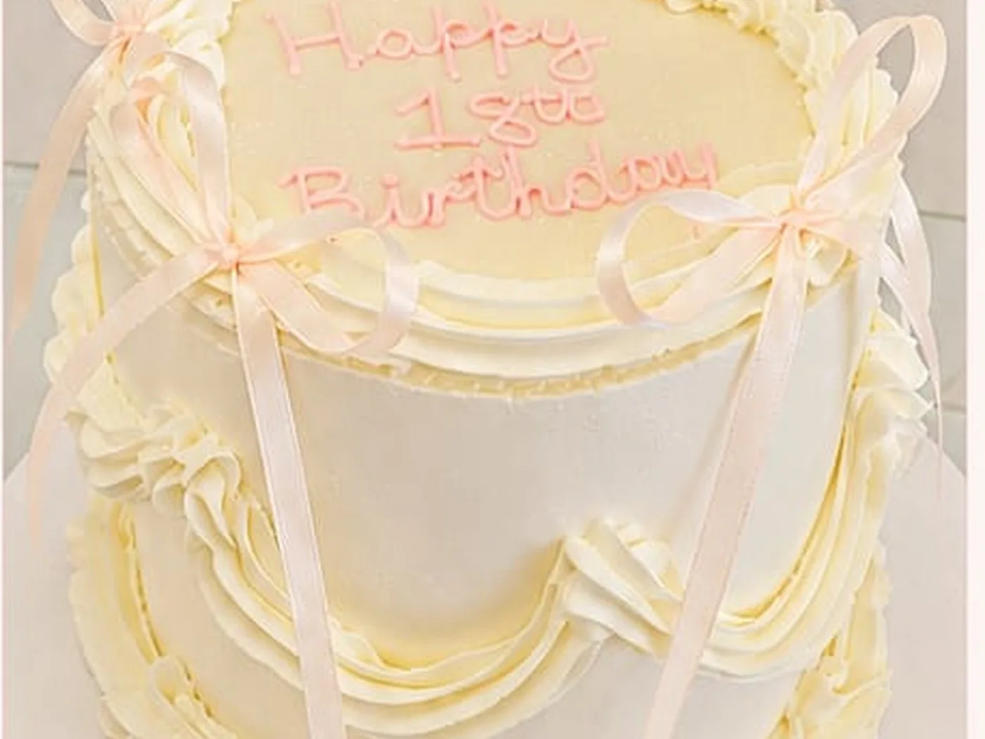 A tall ivory cake piped with buttercream ribbons and bows, with a birthday message written in pink