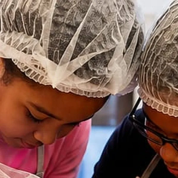 Two children in hairnets and aprons, heads down, concentrating in a baking class